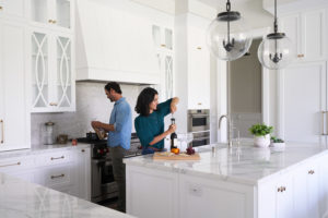 A modern white kitchen with brand-new cabinets. A husband and wife stand in the kitchen making dinner.