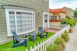 Front yard of a house with a white picket fence. Blue Adirondack chairs sit in front of a large white bay window.